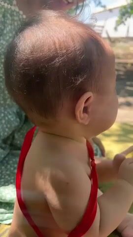 The video shows a baby in the process of crying. The baby is wearing a red ribbon around its neck and a diaper. The baby's face is contorted in distress, with eyes closed and mouth open in a wail. The baby's hands are visible, one holding a small green object.

In the background, a person wearing a green floral dress is partially visible, leaning over the baby. The setting appears to be outdoors, with sunlight filtering through trees, suggesting daytime. The ground is covered with a blanket or mat, and the overall atmosphere is casual and possibly a park or garden. There are no visible text elements in the media. The location context provided is Айдарлы, Kazakhstan. The baby's emotional state is clearly one of unhappiness or discomfort, as indicated by its crying.