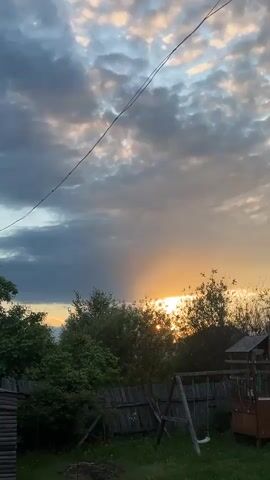 The video presents a static, wide shot of a rural or semi-rural backyard scene at what appears to be either sunset or sunrise. The sky dominates the upper portion of the frame, filled with a mix of grey and light, patchy clouds. A prominent band of golden light from the sun slices vertically through the clouds on the right side, indicating the sun's low position near the horizon. An overhead wire stretches diagonally across the upper part of the sky.

Below the horizon, the landscape features a lush green grassy area in the foreground. A wooden fence runs across the middle ground, behind which numerous green trees and bushes grow, partially obscuring the distant horizon. The sun's rays are visible peeking through the dense foliage on the right side of the frame. To the right, a simple wooden swing set with two swings stands on the grass, next to a small wooden structure, possibly a shed or part of a play area. The overall scene suggests a peaceful and natural setting, likely captured in Новая Тура, Russia. No text is visible in the video.