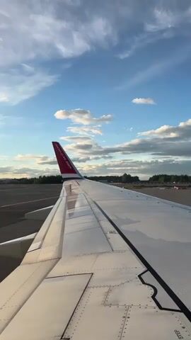 The video begins with a view of an airplane wing with the Norwegian Air logo visible on the tail fin. The wing is white with visible seams and rivets. The sky above is blue with scattered white clouds, and the sun appears to be setting or rising, casting a warm glow. The runway is visible to the left of the wing, with trees and buildings in the distance. Audio includes what appears to be an announcement in a language other than English, followed by the sound of an airplane engine. The announcement states, "We are instructed by the group to perform the jacket from its patch..." The video is short and focuses on the wing and the sky.