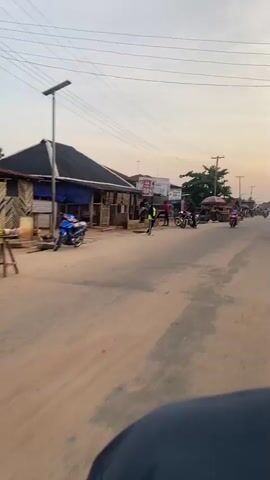 The video captures an eye-level, forward-moving perspective from a vehicle on a paved road in a town or village setting. The road surface is worn, with dirt shoulders and some sand. On the left, a row of single-story buildings with dark roofs and wooden facades lines the street; one building features a dark blue tarp covering its front. Overhead utility wires extend across the scene, supported by poles. A woman wearing a light-colored dress, without a helmet, rides a red step-through motorcycle from the right side of the frame towards the left. In the mid-ground, a person in a bright green top and dark pants stands near a yellow conical object in the middle of the road. Several other individuals are visible standing on the right side of the road, alongside multiple parked or stationary motorcycles. The sky is clear, illuminated by a warm, soft light, indicative of late afternoon or early evening.
