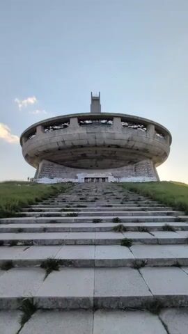 This media file captures the abandoned Buzludzha Monument in Shipka, Bulgaria, under a clear sky, likely during the golden hour as suggested by the soft, warm light illuminating the structure and its surroundings. The monument, a massive, saucer-shaped concrete edifice with a tall central tower, sits atop a grassy hill. A wide, imposing stone staircase leads up to its entrance. The stairs are weathered, with patches of grass growing through the cracks, indicating neglect and the passage of time.

The monument itself appears to be in a state of disrepair. The circular facade has numerous openings, suggesting broken windows or missing panels. A prominent tower rises from the center, also showing signs of decay. White text, likely inscriptions or slogans, is visible on the lower part of the monument's curved wall, though it's too distant and degraded to be legible.

The scene is quiet and somewhat melancholic, evoking a sense of forgotten grandeur and historical significance. The vastness of the structure and its isolated location on the hilltop emphasize its monumental scale and its once important role. The grassy slopes surrounding the monument are bathed in soft light, contrasting with the stark concrete of the building. No people are clearly visible, adding to the feeling of solitude. The weather appears calm and pleasant, with only a few wisps of clouds in the pale blue sky. The overall impression is one of an awe-inspiring, yet eerie, relic of a past era.