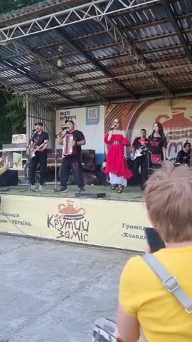 A slightly elevated full shot shows an outdoor stage with five musicians performing. A female singer in a red dress is at the center, flanked by two men, one playing an accordion and the other a guitar. Two other musicians, one male and one female, play guitars on the right. In the foreground, spectators are seen from behind, including a woman in a yellow shirt with a gray backpack, holding a phone. Another person in a yellow shirt is closer to the stage on the left, also recording. The stage has a yellow banner with Ukrainian text and a QR code. Trees are visible in the background.