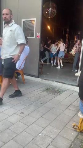 The video captures a lively scene unfolding in front of an open doorway, possibly a bar or event space, in what appears to be an outdoor patio area. The foreground shows a paved courtyard with scattered patches of grass. The midground features a group of people engaged in dancing and socializing.

The subjects include several individuals, men and women, dressed in casual attire. One man in a white t-shirt and blue jeans walks into the frame from the right, carrying a plate. Others are captured mid-dance, with dynamic movements and expressions suggesting enjoyment. The setting is urban, with a building entrance featuring a red security camera sign with the word "DIRECT" visible. Inside the building, a disco ball hangs from the ceiling, reflecting light, and stage lighting illuminates the interior. Furniture, possibly lounge seating, is visible in the background.

The activity is clearly a social gathering with dancing, likely a party or event. The time of day appears to be late afternoon or early evening, given the lighting conditions and the overall atmosphere. The weather seems pleasant, as people are comfortably dressed in short sleeves and shorts. The location is suggested to be Karlstad, Sweden, though this is not explicitly visible in the footage itself. The emotions conveyed are positive, with a sense of energy and fun.