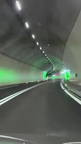 The video shows a point-of-view shot from inside a modern road tunnel, likely in Zurzach, Switzerland, as a vehicle travels forward. The tunnel features a smoothly paved asphalt road with clear, double white solid lines marking the center. The walls of the tunnel are a mix of light grey concrete and possibly reflective panels, illuminated by bright white LED lights arranged in a line along the ceiling. A distinctive feature is the prominent green accent lighting, which washes sections of the tunnel walls and highlights various architectural elements and emergency exits along the route. As the vehicle moves, the tunnel appears to curve gently to the right in the distance. No other vehicles or people are visible, and there is no discernible text. The overall impression is one of a clean, well-maintained, and technologically advanced infrastructure.
