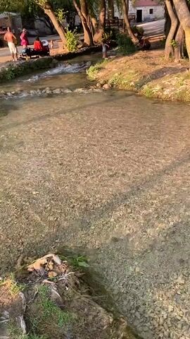 The video opens with a wide shot of a shallow, clear river flowing over a gravel bed. The sun is shining, casting dappled shadows on the water from the trees on the left bank. In the background, on the left bank, several people are visible, some sitting and some standing. On the right bank, more trees and vegetation line the river. As the video progresses, the camera pans slowly to the right, following the flow of the river. The water appears to be shallow enough to wade through in many areas. Towards the end of the video, the camera focuses on a small section of the river where a makeshift dam of rocks has been constructed, causing the water to ripple and create a small cascade. The sun continues to cast long shadows, indicating it is likely late afternoon or early evening. The overall impression is one of a tranquil, natural setting, possibly a park or a recreational area near a river.