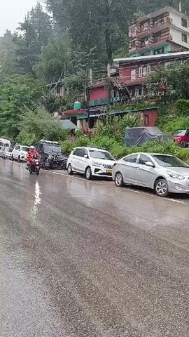 A motorcycle rider wearing an orange raincoat and a white helmet is moving to the right on a wet, asphalt road. Multiple cars are parked on the left side of the road. Behind the parked cars, there are buildings with red and brown accents built on a hillside. To the right of the road, there is a bus and other vehicles. The sky is overcast.