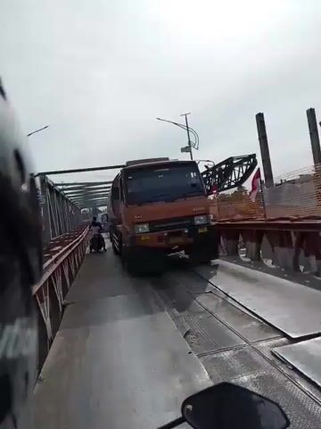 A dashcam recording shows a semi-trailer truck with a tanker body traveling across a bridge. The truck is orange and white with a "6X2" marking on the front. The truck appears to be driving on the right side of the road. In the background, other vehicles and people can be seen. The bridge has metal railings on the sides. The sky is overcast.