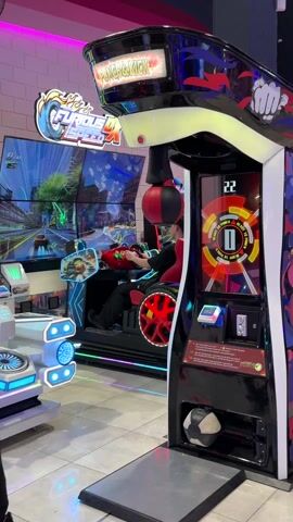 Here is a description of the video:
The video shows a brightly lit arcade. The focus is on a "Punch & Kick" arcade game. A man is standing in front of the game, throwing punches at a hanging punching bag.  The game tallies points on a digital display, showing numbers like 1058, 2063, and 3029. Another arcade game, "Cruis'n Blast," is visible in the background, and a young man is sitting in it, playing. The "Punch & Kick" game has a colorful design, including images of a fist, flames, and geometric patterns.  Other arcade games and lighting are also present in the background, creating a lively arcade atmosphere.