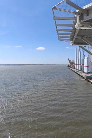 The video shows a wide body of water under a clear blue sky with a few scattered clouds. In the foreground, the water has small waves. On the right side of the frame, there is a dock or pier with large cylindrical objects, possibly wind turbine components, stacked vertically and secured with straps. A yellow crane is also visible on the dock. In the distance, on the water, there are several small boats moving. Part of an elevated metal structure with a safety net is visible in the upper right corner of the frame.