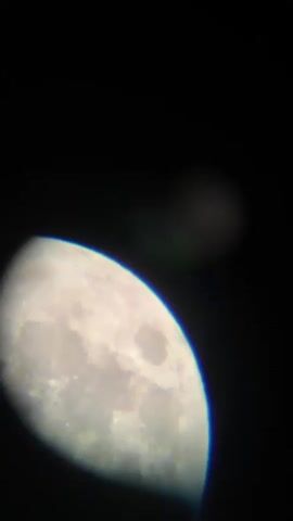 This media file shows a close-up, slightly blurry, view of the moon against a black sky. The moon is illuminated and its craters are visible, indicating it is likely nighttime. In the background, there is a faint, out-of-focus object, which could be another celestial body or a light source. The overall impression is that of an astronomical observation, possibly taken with a telescope. There is no visible text in the media file. The location context of Târgu Neamț, Romania, suggests this image was captured from that region, but there are no specific visual cues within the image to confirm this. The activity depicted is the observation of the moon. The time of day is clearly night. There is no information about weather or emotions.