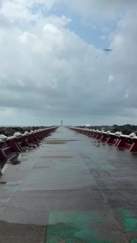 The video shows a wet concrete pier extending into the sea under a cloudy sky. The pier is lined on both sides with rocks and concrete barriers painted red and white. In the distance, at the end of the pier, is a structure resembling a lighthouse or tall tower. Several birds are flying in the sky above the pier. The pier is wet, reflecting the sky and surrounding elements.