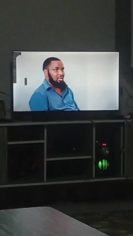 The video shows a television screen displaying two people, alternating between them in quick succession. The first person is a woman with dark skin, wearing a white headscarf and an orange top. The second person is a man with dark skin, a trimmed beard, and a blue shirt. The television is placed on a grey and white entertainment center. To the right of the television, on the bottom shelf of the entertainment center, is a computer tower with green lights visible on the front panel. A small portion of a grey table is visible in the foreground. The environment appears to be a dimly lit room.