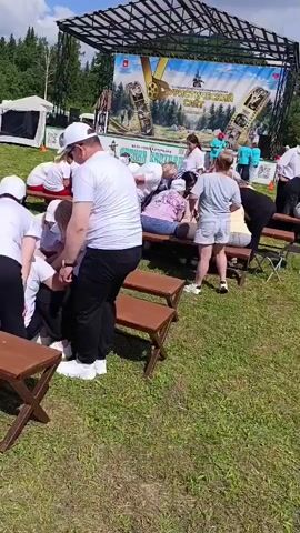 A group of people are gathered outdoors on a sunny day, with a stage set up in the background. Most of the individuals are seated on benches, facing inward towards a central point. They appear to be participating in an activity or game. One person in the foreground, facing away from the camera, is wearing a white hat and a white t-shirt. The overall atmosphere suggests a community event or festival.
