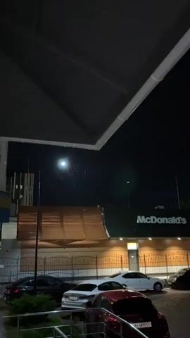 This is a vertical, low-light video shot at night, likely during a light rain. The scene is a McDonald's restaurant in Hatne, Ukraine. The iconic golden arches logo is visible on the green facade of the building, illuminated by warm yellow lights. The interior of the restaurant is visible through large windows, showing a well-lit space with patrons.

In the foreground, several cars are parked in a lot. These include a dark sedan on the left, a white sedan, a red hatchback with a visible license plate, and a dark-colored van or SUV. A white trailer is also parked in the lot. The cars and the McDonald's lot appear to be on a paved surface.

The sky is dark, indicating nighttime. A faint light, possibly the moon or a distant streetlight, is visible in the upper left quadrant of the frame. Streaks of light are visible in the sky, suggesting rain is falling, creating a slightly blurry effect. The overall mood is quiet and somewhat serene, despite the commercial setting.

Notable text visible includes "McDonald's" on the building's signage. The license plate on the red car appears to read "AI 4890 CO." The time of day is clearly night, and the weather is rainy. The setting is a fast-food restaurant in Ukraine.