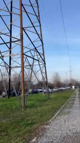 The video shows a person walking away from the camera on a gravel path. To the left is a large metal electrical tower. Next to the path is a grassy area and a road with a line of cars. In the distance, more electrical towers can be seen against a blue sky with some clouds. The person is wearing a dark coat and has long dark hair. The path is lined with green grass on one side and a row of cars parked along the road on the other.