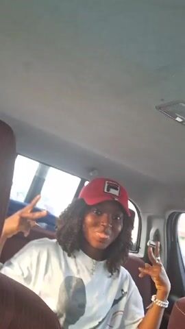 The video depicts a young woman wearing a white t-shirt with a graphic print and a red baseball cap with a logo on it. She has curly hair and is wearing a necklace with a cross pendant. She is seated in the back of a car with maroon-colored seats. The car is in motion, and there is a window visible in the background. The interior of the car is well-lit. The woman initially sticks her tongue out and then smiles at the camera. An arm appears briefly in the frame near the beginning of the video.