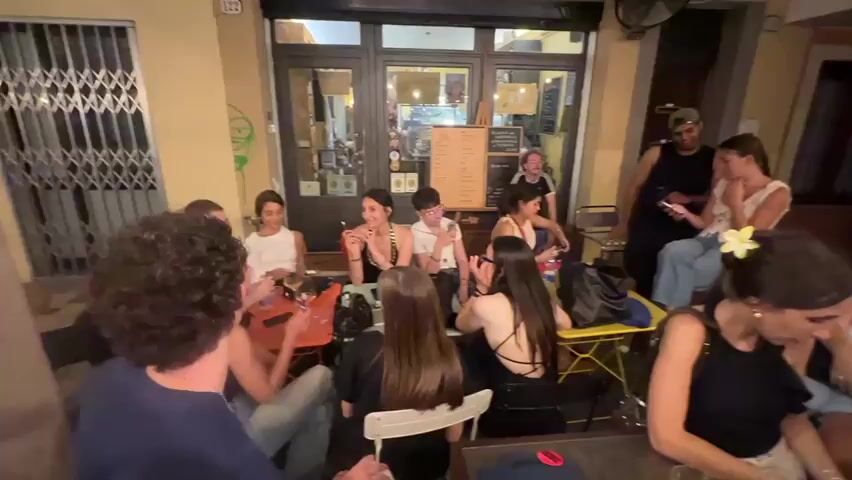 The video shows a gathering of people sitting and standing outside what appears to be a cafe or restaurant. The scene is dimly lit, suggesting it is evening or night. Several tables and chairs are visible, with some people seated and others standing. Many individuals are looking at their phones, and some are engaged in conversation. The overall atmosphere seems relaxed and social, with people socializing and enjoying themselves in an outdoor setting.