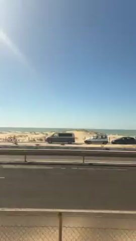 The video shows a view from an elevated position overlooking a two-lane road that runs alongside a beach. The beach consists of sand dunes leading to the ocean. There are several parked vehicles on the beach side of the road, including vans and cars. Bicycles are leaning against the low dune fence. The sky is blue with the sun shining brightly, slightly obscuring the view at the top. The perspective suggests the video was taken from a building or structure next to the road. The camera moves slightly, panning along the road and beach.