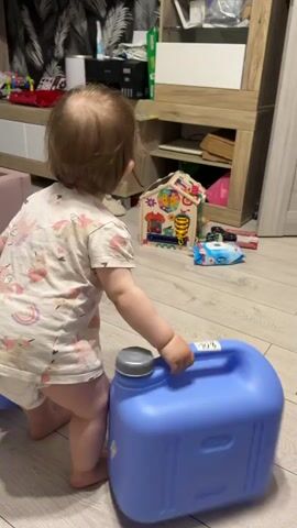 Here's a detailed description of the video:

A young child, likely a toddler, is seen from the back, carrying two matching blue plastic containers, one in each hand. The child wears a light-colored top with a unicorn and rainbow pattern, and what appears to be a diaper. They are barefoot.

The background shows a cluttered living space. A light pink loveseat is visible to the left. A light wood-toned cabinet or entertainment center is behind the child, holding various items. Several toys are scattered on the floor, including a toy house, a small blue object with white polka dots, and other miscellaneous items. The flooring is light wood-grain. A black and white patterned curtain or tapestry hangs on the wall.