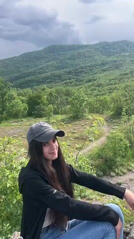 A young woman with dark hair is sitting in a grassy area with shrubs and small trees, with a distant, wooded mountain in the background. She is wearing a dark-colored jacket, jeans, and a gray baseball cap. She points up towards the mountain, and then towards the sky. The sky is overcast with dark clouds. The overall tone of the video is peaceful and natural.