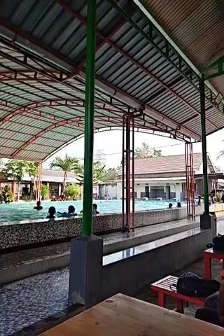 The video shows a swimming pool area with a covered roof. Several people are in the pool, some appear to be swimming and others are standing or sitting in the water. There are palm trees and buildings visible in the background. In the foreground, there are tables and chairs, suggesting an area for relaxation or dining. The overall atmosphere appears to be a recreational setting.