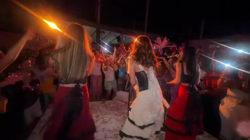 The video captures a lively night-time event with a focus on three women dancing on a raised platform. They are viewed from behind, wearing elaborate long skirts: one red and black, one white with black accents, and another red and black. The woman in the white dress is in the center. The surrounding crowd, blurred in the background, is cheering, raising their hands, and appears to be enjoying the performance. The scene is illuminated by warm orange and purple lights, creating a festive atmosphere.