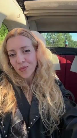 A woman with long blonde curly hair, wearing a black leather jacket and a scarf, smiles at the camera. She is sitting in the passenger seat of a convertible car. The top of the car is down, and the sky and some greenery are visible in the background. The woman's seatbelt is fastened. The lighting suggests it is daytime, possibly late afternoon given the warm tones on her hair. The interior of the car shows the back seats with red and white upholstery.