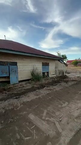A wooden building with a red tin roof stands on stilts. The building has two blue shuttered windows and a blue door. The area around the building is covered in mud and debris, suggesting recent flooding or construction. The sky above is cloudy with patches of blue. In the background, more buildings with similar architecture and red tin roofs can be seen. The camera pans slightly to the left, revealing more of the muddy terrain and the surrounding structures.