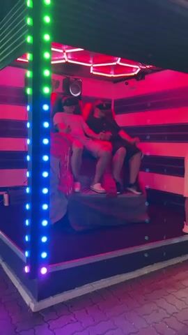 Two young men wearing VR headsets are seated in a simulated ride or attraction. The attraction features a red, sculpted seat and is enclosed within a structure adorned with horizontal black and pink stripes. The environment is further enhanced by colorful, pulsating LED lights, primarily green and pink, that illuminate the scene. One man on the left is wearing a light-colored t-shirt and shorts, while the man on the right is wearing a dark t-shirt and shorts. Both are holding controllers, suggesting they are actively engaged in a virtual reality experience. The overall setting appears to be an entertainment venue or arcade, possibly outdoors given the visible pavement and the way the structure is set up. The lighting creates a dynamic and immersive atmosphere.