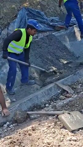 Three construction workers are pouring and spreading wet concrete using shovels. The workers are wearing high-visibility vests over their clothing. One worker in a blue cap and yellow vest is on the left, using a shovel to move concrete into a channel. Another worker in a green cap and dark jacket is in the center, also using a shovel to work the concrete. A third worker, partially visible in the background on the right, is wearing a yellow vest and blue pants and is also using a shovel. The area appears to be an outdoor construction site with dirt, gravel, and a blue tarp visible in the background. Several tools, including shovels and what appear to be concrete finishing tools, are lying on the ground.