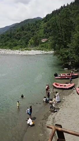 A wide shot shows a turquoise river with a sandy bank on the right. A group of people, dressed in casual summer clothing, are gathered on the bank and wading in the shallow water. Some are standing, others are sitting or lying down. Two red inflatable rafts are visible on the bank. In the background, a lush green forest covers the rolling hills, with a few scattered buildings visible amongst the trees. The sky is partly cloudy. Several individuals are engaged in activities in the water, suggesting a recreational setting.