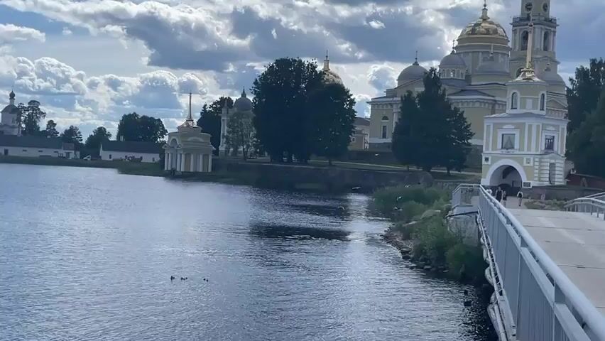 A pedestrian bridge spans a body of water, leading towards a large, yellow, domed building complex. The complex appears to be a monastery or church, with multiple golden domes and spires. Trees are situated behind the building. Several people are walking across the bridge. The sky is cloudy, with patches of blue visible. The water is calm with a few ducks on its surface.