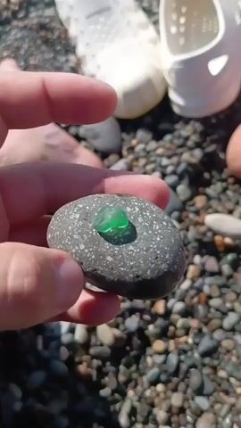 A hand is holding a smooth, dark gray, oval-shaped rock. A small, irregularly shaped piece of translucent green sea glass is resting on top of the rock. The background consists of many small, dark-colored pebbles, indicating a beach or rocky shore. In the upper background, out of focus, are parts of two white objects, possibly footwear or plastic containers. The lighting suggests it is daytime, with the sun casting a shadow from the sea glass onto the rock.