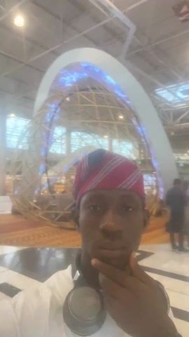 A person with dark skin is taking a selfie in what appears to be a large, public space such as an airport or convention center. The person is wearing a white shirt, a patterned red and white head covering, and black headphones are around their neck. They are holding their hand to their chin in a thoughtful pose. In the background, there are other people, large windows, and elements of the building's architecture like ceiling beams and lights. The overall lighting is bright and airy.