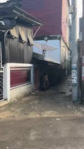 The video pans across a street scene with a mix of buildings and some parked motorcycles. In the foreground, a red scooter is parked with a person wearing a red helmet sitting on it. To the right of the scooter, there are several large bags, possibly filled with goods, next to a commercial freezer displaying an ice cream logo. Further down the street, food stalls are visible with signage in Indonesian. One stall displays "MARZABAK BOMBAY" and prices, while another has "ADA NASI UDUK". A small shop with a maroon-colored storefront and a sign that includes the word "CONSEL" is also visible. The environment appears to be a commercial area with a somewhat worn and weathered appearance, suggesting a busy street with ongoing commerce.