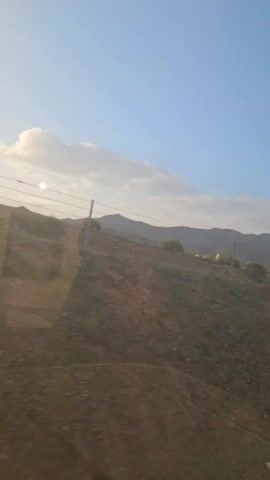 The video shows a view of a landscape from a moving vehicle, possibly a train. The scenery includes fields, some appearing green, and distant mountains under a blue sky with a few clouds. There are also signs of human presence such as a fence and some abandoned construction-related items by the roadside. The video is shot during daylight with the sun visible through the clouds. The camera's view is partly obstructed by the window of the vehicle.