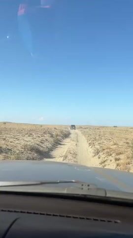 The video shows a view from inside a vehicle, looking out through the windshield at a dirt road. The road is dry and dusty, with tire tracks visible. The surrounding landscape is arid and appears to be a desert or plains area, with sparse vegetation and sand dunes. The sky is clear and blue, with a few faint clouds. In the distance, another vehicle, a dark-colored SUV, is driving on the same dirt road. The camera is focused on the road ahead and the other vehicle. The video appears to capture a moment during an off-road excursion or journey.