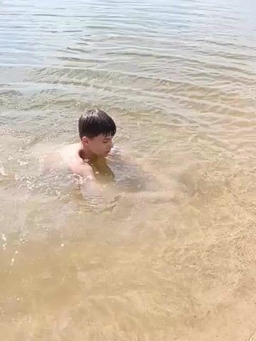 A young person is swimming in shallow, murky water. The person is on their stomach, with their head turned to the left, and their arms are outstretched in front of them. The water is clear enough to see the sandy bottom, and there are ripples on the surface. The person appears to be startled by something in the water, as they exclaim "Someone is biting me."