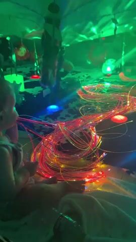 A young child sits on a blanket and plays with a bundle of fiber optic lights in a dimly lit room. The lights are multicolored and coiled together. The child holds a few strands and appears to be putting them in their mouth. In the background, green lights project patterns onto a fabric ceiling, and other lights, some red and blue, are visible. Two other figures, possibly children, are blurred in the background, moving. A plastic bottle sits on the blanket near the child.