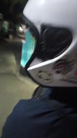 The video shows a first-person perspective of someone wearing a white helmet with stickers and a dark blue jacket. The person appears to be riding a motorcycle or scooter at night. The helmet has several stickers, including a panda, a shark-like graphic, and a digital clock displaying "07:35". The surrounding environment is dimly lit, with hints of buildings and streetlights. The video captures the motion of the rider as they navigate the road.