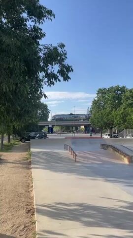 The video captures an outdoor skatepark in Santa Perpètua de Mogoda, Spain, on a sunny day with a clear blue sky. The scene is framed by lush green trees on either side. In the background, an elevated train track with a bridge spans across the landscape.

As the video begins, a modern train crosses the bridge. Below the tracks, a street is visible with pedestrians and a few cars. The camera then focuses on the skatepark itself, showing concrete ramps and metal grind rails. A young male skateboarder, wearing a green t-shirt and grey pants, approaches one of the grind rails. He successfully performs a rail grind, sliding along the metal bar on his skateboard.

Throughout the video, various people can be seen walking in the background. Graffiti, with the word "GRAFFITI" partially visible, is present on one of the concrete ramps within the skatepark. The setting appears to be a vibrant public space during daylight hours.