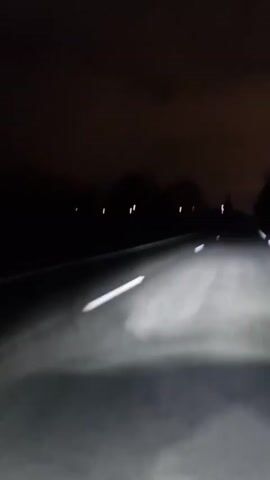 A nighttime view from inside a vehicle moving along a paved road. The road is dark gray or black, illuminated by the vehicle's headlights. White lane markings are visible. The sky is dark with a hint of cloud cover. Trees and sparse lights can be seen in the distance on either side of the road, adding to the darkness. The camera appears to be mounted on the dashboard or windshield, capturing the forward perspective of the driver. The road appears to be largely empty and the car seems to be the only vehicle traveling along it.