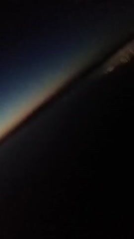 The video depicts a dimly lit scene with a gradient sky visible at the top. The sky transitions from a dark blue at the top to a lighter orange near the horizon. A dark expanse, likely water, occupies most of the frame. In the distance, a faint line of lights suggests a city or coastline. The perspective is from an elevated position, possibly from an airplane or a tall building. The camera is tilted slightly.