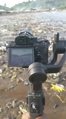 Here is a concise description of the video:

The video shows a camera with an attached gimbal pointing towards a beach covered in trash. The beach is muddy, and there are various pieces of plastic waste, bags, and other debris scattered across it. In the distance, there is a coastline with some buildings and people walking along the shore. The shot is static.
