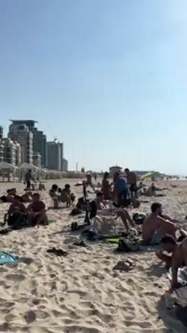 The video shows a sandy beach on a sunny day, filled with people. Many are sunbathing, either sitting or lying down, while others are walking or standing. In the foreground, a woman in a red bikini stands next to a man in a blue shirt and straw hat, who appears to be selling something from a cart he's pushing. Another shirtless man stands nearby. In the background, tall buildings and construction cranes are visible on the horizon, indicating an urban environment. A lifeguard stand can also be seen behind the group.