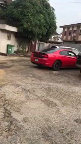 The video shows a red Dodge Charger parked in a concrete lot. The car has black rims, a black spoiler, and dual exhaust pipes. A black stripe runs along the side. The license plate is green. There is a green trash can and a tree to the left of the car. A white SUV is partially visible behind the Charger. A house is visible in the background. The ground is grey and concrete.