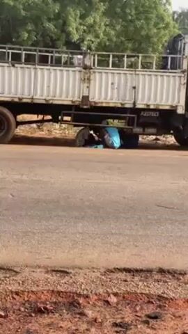 The video shows two people kneeling down under the body of a large white truck on the side of a road. The person on the left is wearing a striped shirt and baseball cap. The person on the right is wearing a light blue sleeveless shirt and light-colored shorts, with a baseball cap. Their backs are to the camera, and they appear to be working on something placed between them on the ground. There are bags and items on the ground near them. The road is asphalt, and the surrounding area has reddish soil and green trees.