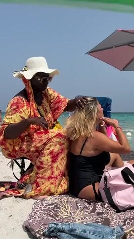 A dark-skinned person wearing a yellow and red tie-dye robe and a white hat is sitting on a beach chair and brushing the hair of a light-skinned person wearing a black swimsuit. The light-skinned person is sitting on a patterned blanket and facing away from the camera, looking to their right. The background shows a beach with sand and a body of turquoise water. There is a beach umbrella in the upper right corner. A pink bag is on the blanket to the right of the light-skinned person.