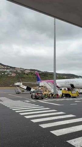 The video presents a view of an airport tarmac under a cloudy sky. A Wizz Air airplane is the main focus, positioned to the left, and a Ryanair plane is seen to its right, both connected to mobile staircases for passenger boarding/deboarding. Ground support vehicles, including a tug and a small service vehicle, are adjacent to the Wizz Air plane. A crosswalk is visible in the foreground, leading towards the aircraft. The background reveals a hillside dotted with residential buildings.