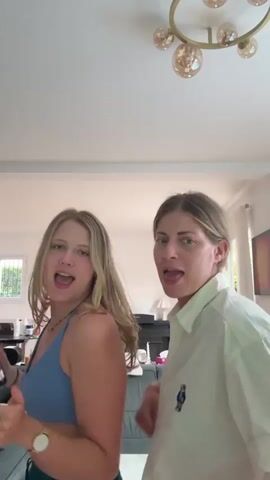 Two young women are dancing. The woman on the left has blonde hair and is wearing a blue bikini top and green athletic shorts. She has a watch on her left wrist. The woman on the right has blonde hair pulled back and is wearing a white collared shirt. They are in a room with a couch, a television, and a chandelier. The lighting is bright.